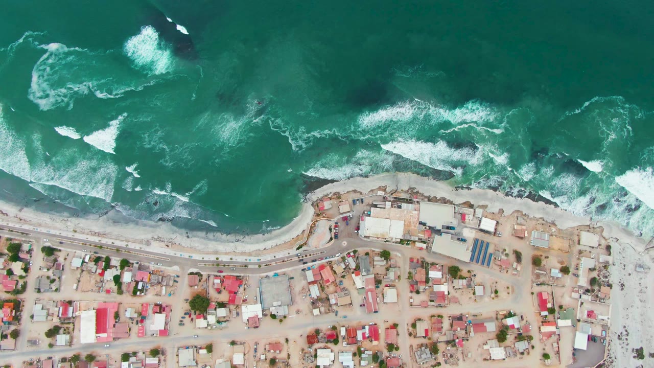 Overhead shoreline with white foam bands, beach, road, and clustered buildings.