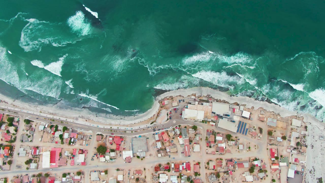 Overhead view of Punta Abreojos with turquoise and dark green water, broken surf near the sandy beach, and a strip of village streets and buildings below, no visible people.