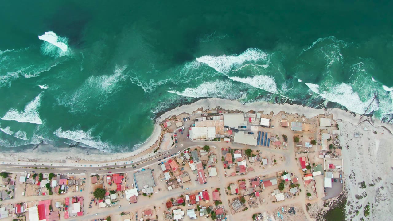 High aerial frame over Punta Abreojos, Mexico, with green-blue ocean and white surf above a shoreline road and compact village rooftops, no visible people.