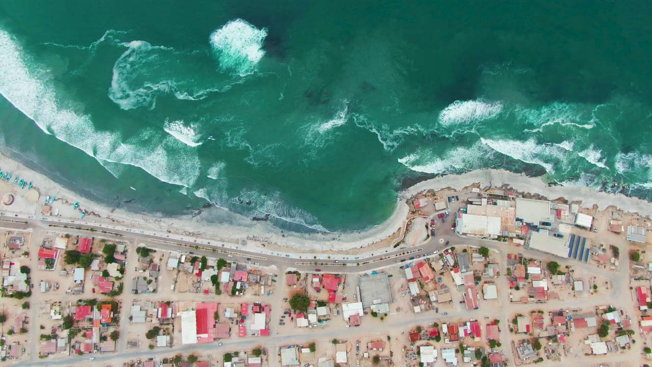 Top-down aerial frame of Punta Abreojos, Baja California Sur, Mexico, with turquoise ocean, white breaking surf, a sandy beach, beach road, compact houses, and no visible people.