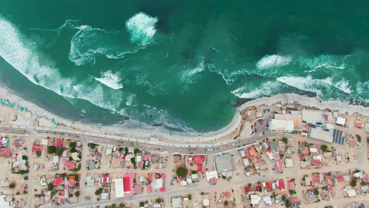 Aerial overhead frame of Punta Abreojos with rolling white waves near the beach, turquoise water across the top, road and residential roofs below, and no visible people.
