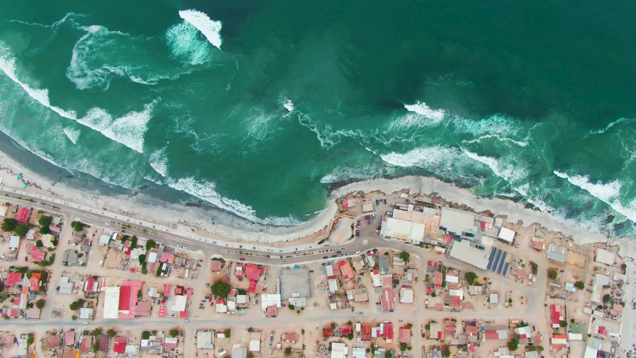 Overhead view of Punta Abreojos with turquoise surf breaking above a pale beach, a road, sandy lots, red-roofed buildings, and no visible people.