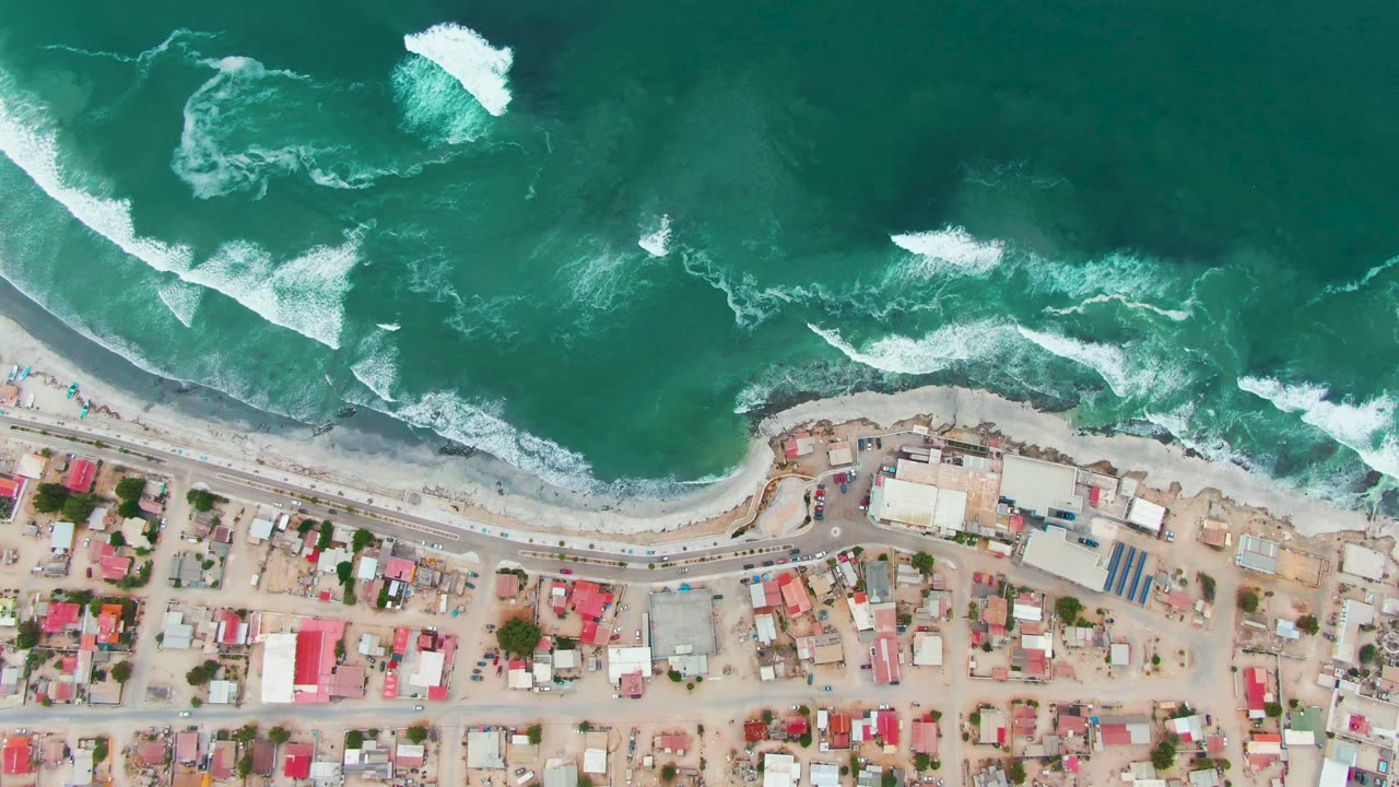 Top-down view of Punta Abreojos with white surf bands crossing green-blue water, a narrow beach margin, roadside buildings, red roofs, and no visible people.