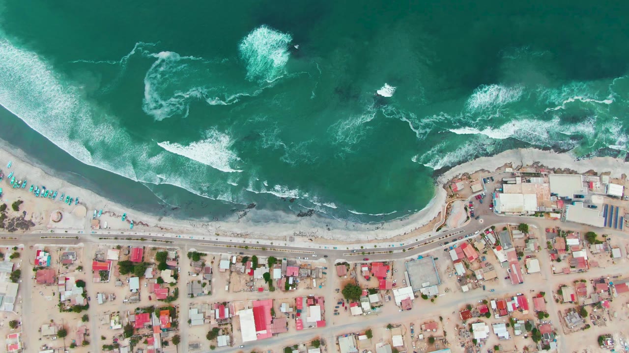 Top-down aerial frame over Punta Abreojos showing turquoise surf above a sandy beach, a road parallel to shore, clustered houses below, and no visible people.