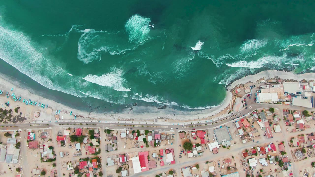Aerial frame over Punta Abreojos with foamy patches offshore, a narrow sandy beach with turquoise umbrellas, a shore road, and low residential buildings with no visible people.