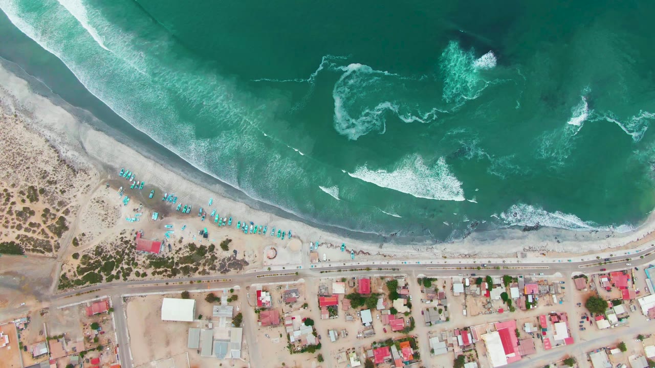 Top-down view of Punta Abreojos with turquoise surf breaking onto a sandy beach, blue umbrellas along the shore, and coastal buildings below the road, with no visible people.