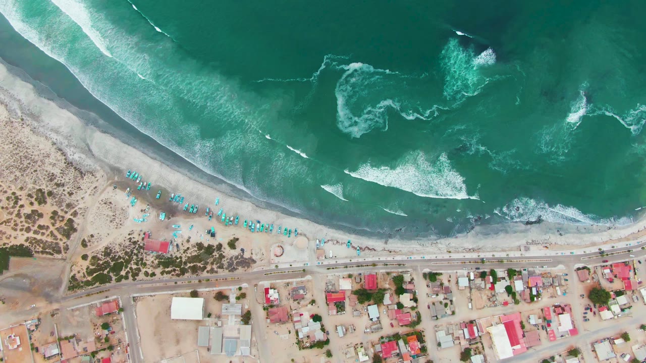 Aerial view of waves advancing toward a beach with umbrellas and buildings.