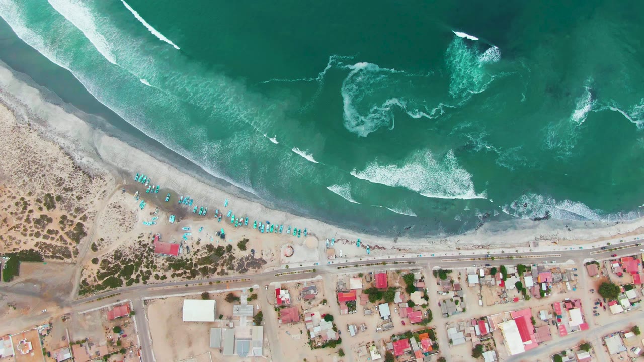 The Punta Abreojos shoreline remains in an overhead layout as foamy surf patterns spread across the green water above the sandy beach and roadside buildings, with no visible people.