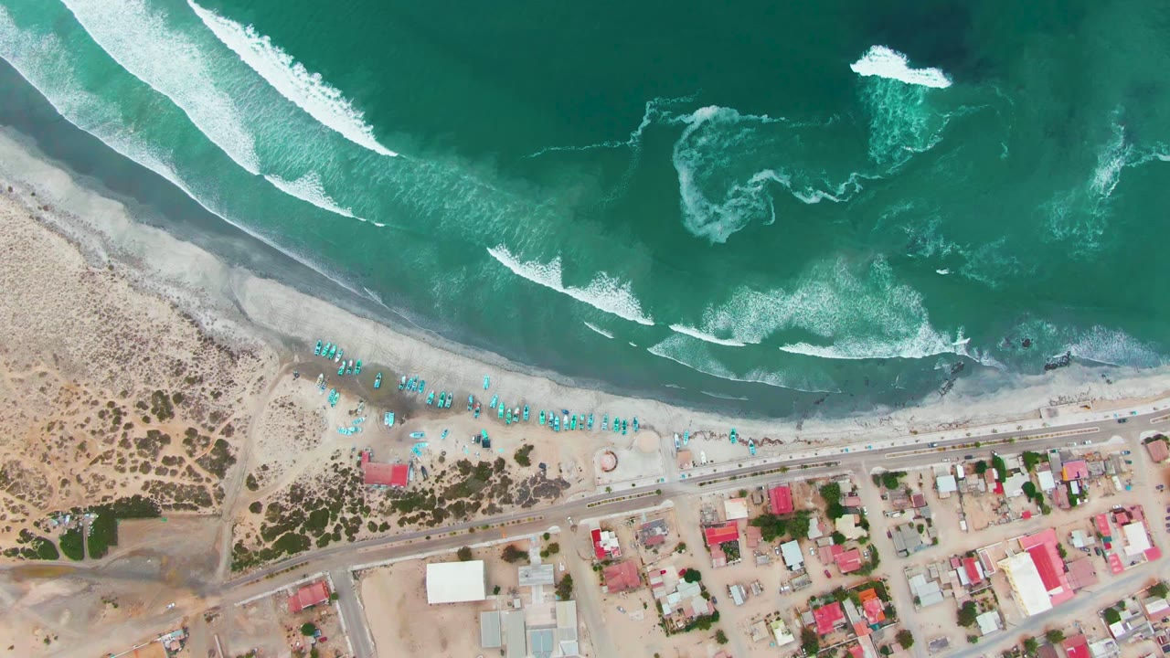 Overhead view of Punta Abreojos shoreline with turquoise surf breaking toward a sandy beach lined with blue umbrellas, a road, and low coastal buildings, with no visible people.