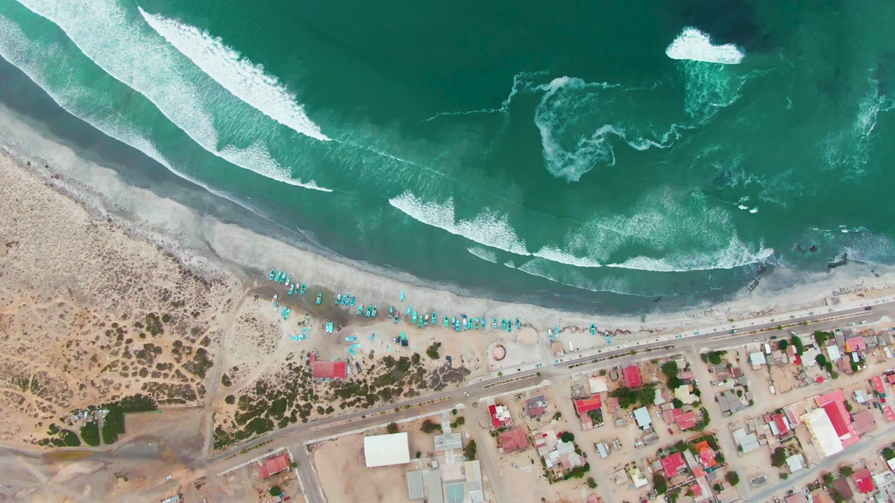 Top-down view of Punta Abreojos coastline where white foam patterns spread across green-blue water above the sandy beach, umbrella line, road, and town buildings, with no visible people.