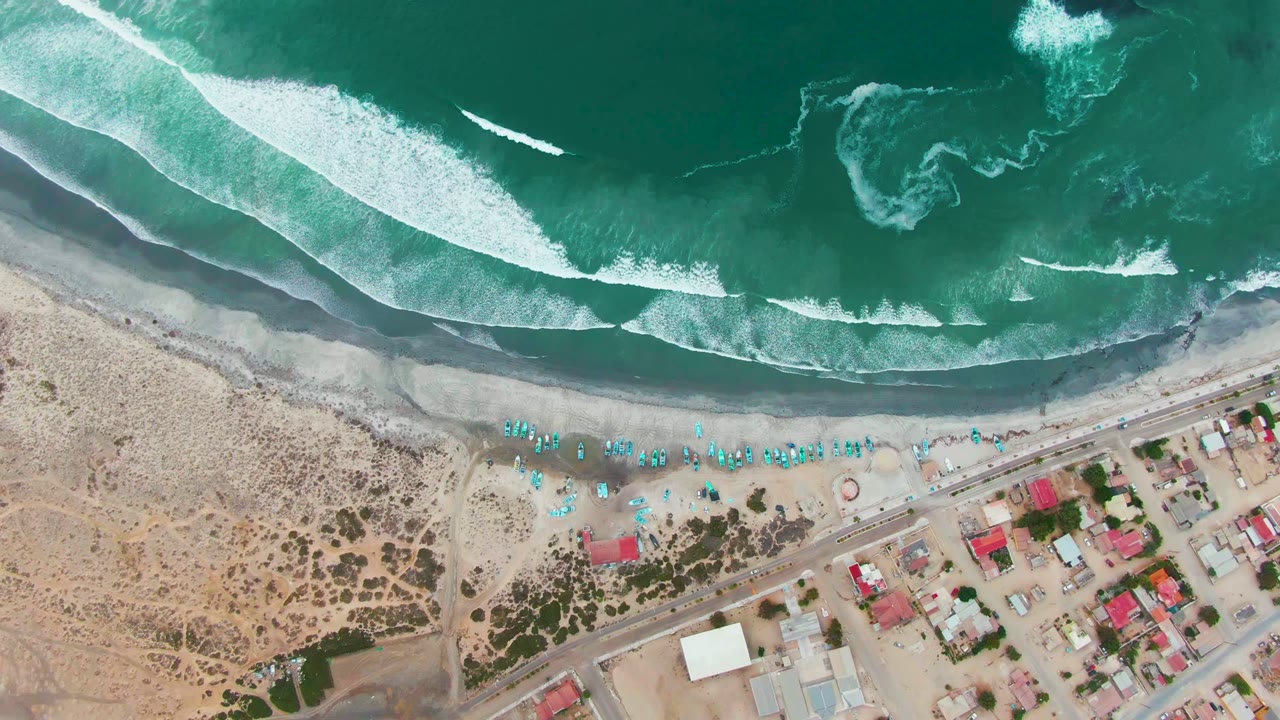 Overhead view of Punta Abreojos surf breaking toward a sandy beach with blue chairs, a shoreline road, low buildings, and no visible people.