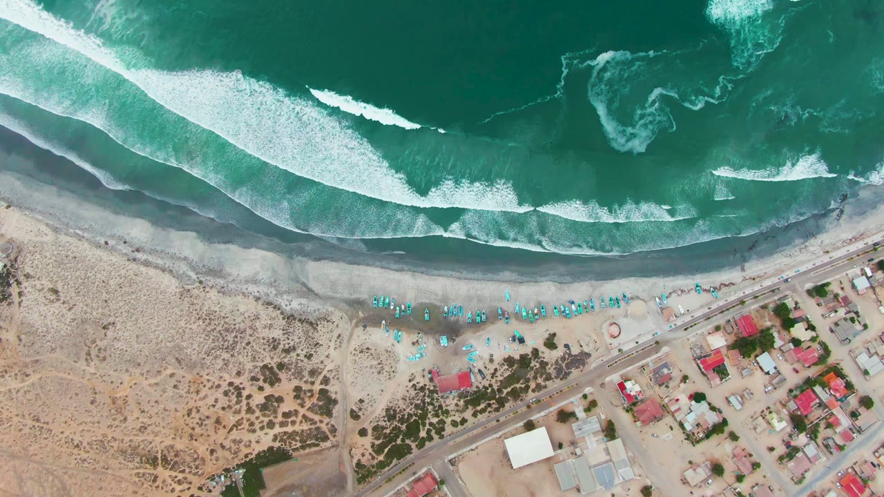 Top-down view in Punta Abreojos with white surf curling across dark wet sand, rows of blue beach chairs, a coastal road, buildings, and no visible people.
