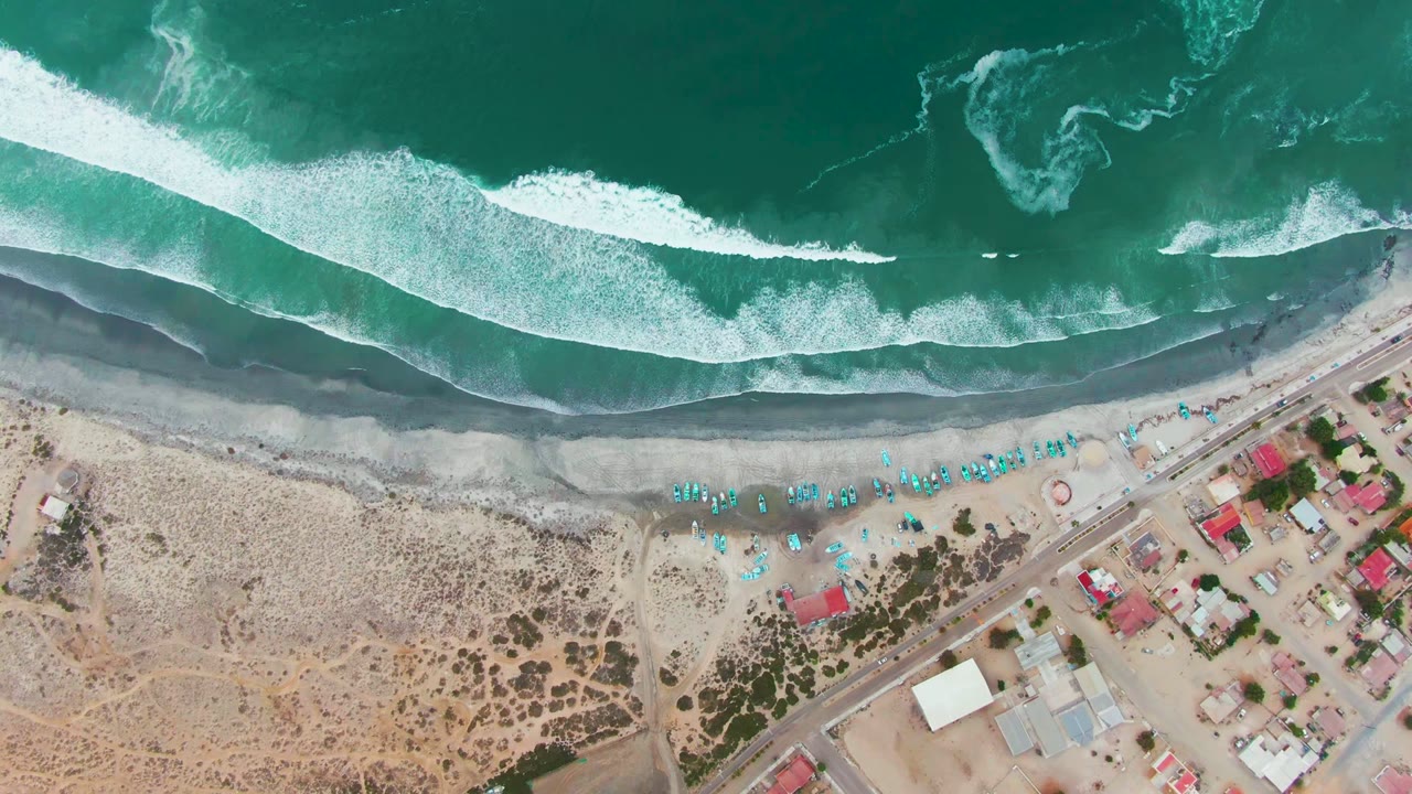 Overhead view of Punta Abreojos, Baja California Sur, with turquoise surf breaking onto a narrow dark shoreline, blue beach umbrellas, small huts, road, and dry sand below, no visible people.