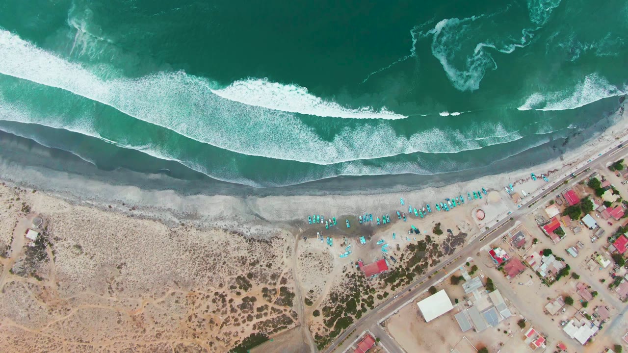 Aerial coastline with white waves, dark shore, blue umbrellas, and beach buildings.