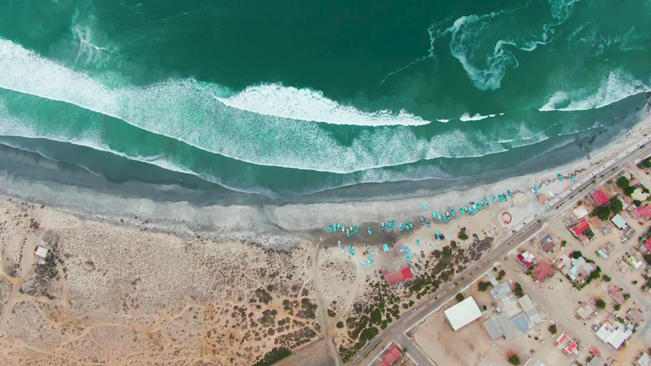 Top-down Punta Abreojos beach view with a long white wave band pressing toward dark wet sand, blue umbrellas near the waterline, huts and a diagonal road below, and no visible people.