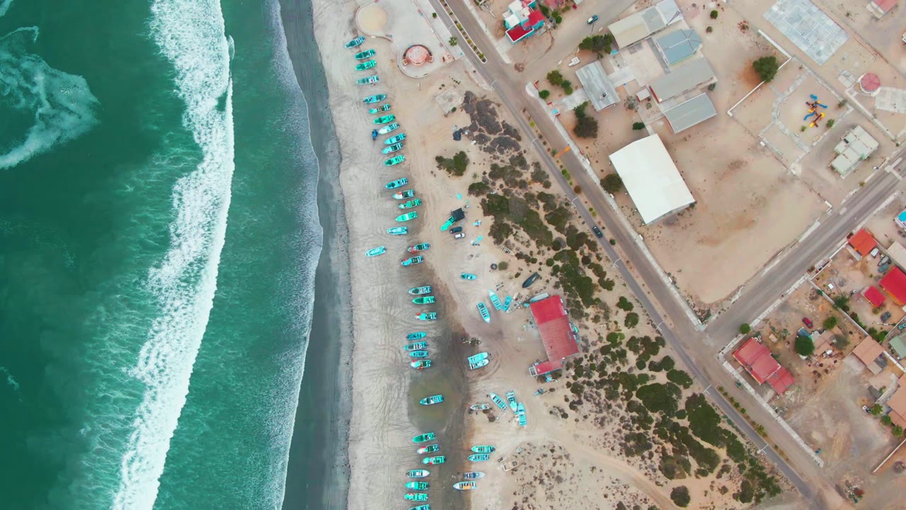 Top-down aerial frame over Punta Abreojos, Baja California Sur, with turquoise ocean surf on the left, pale sand and rows of small turquoise boats or umbrellas near the shore, inland roads and low buildings to the right, and no visible people.