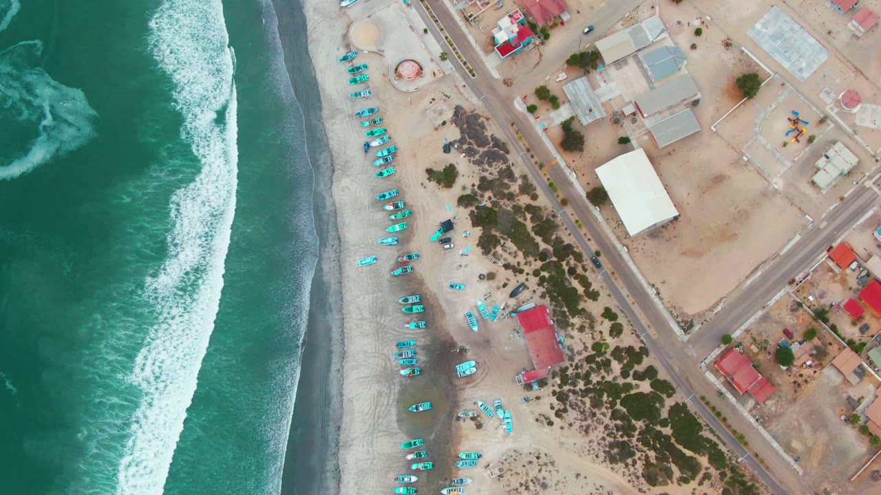 Aerial coastline with breaking surf beside sand, turquoise beach items, roadways, and buildings.