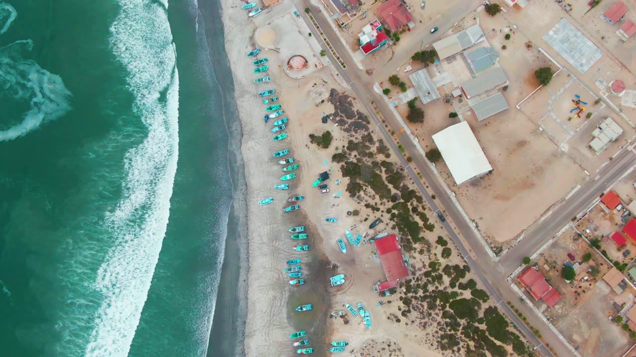 Top-down aerial frame in Punta Abreojos with the surf line shifted along the left edge, pale beach and turquoise shore objects in the center, roads and sparse coastal buildings on the right, and no visible people.