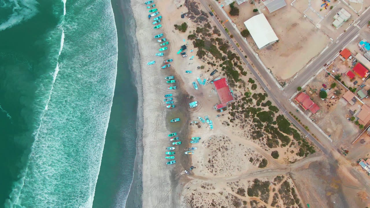 Overhead view of Punta Abreojos with turquoise surf at left, a sandy beach lined with blue beach gear, roads and low buildings inland, and no visible people.