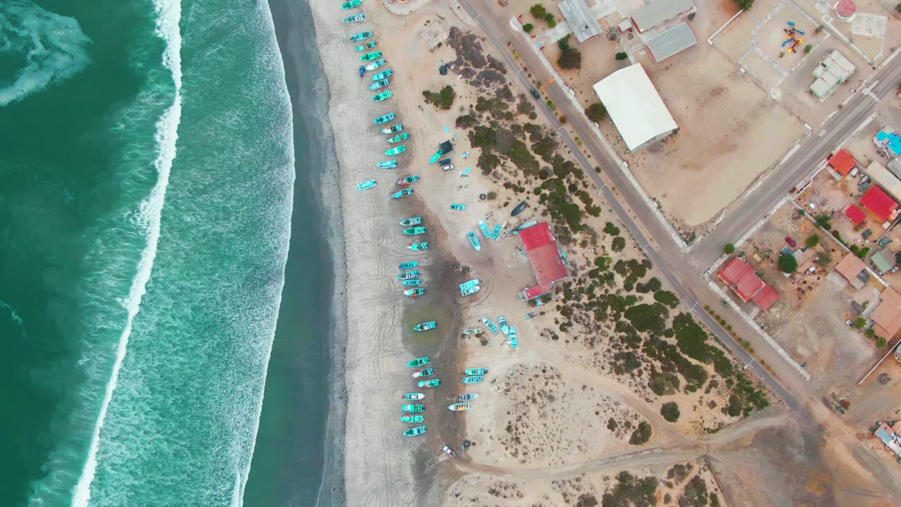 Overhead Punta Abreojos beach frame with turquoise water and white foam on the left, blue beach gear arranged on the sand, coastal buildings and roads on the right, and no visible people.