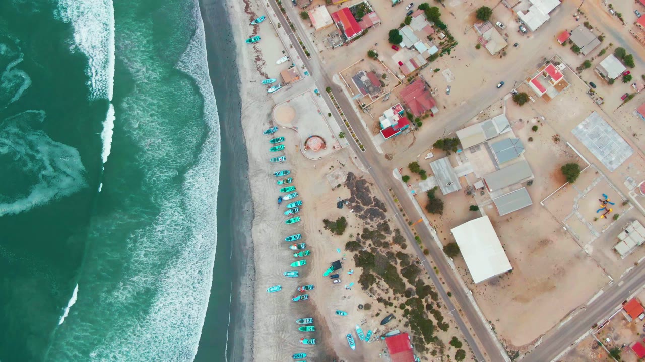 Overhead drone frame of Punta Abreojos with green ocean waves on the left, a sandy beach lined with blue umbrellas, a road, low buildings, and no visible people.