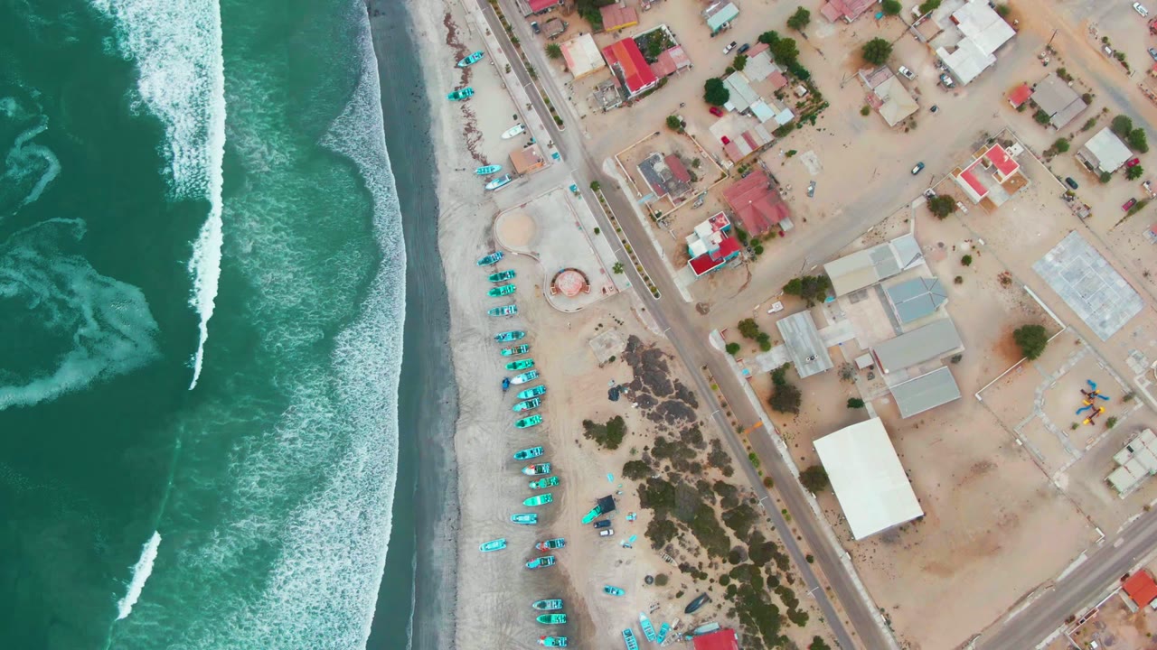 Aerial beach view with breaking waves beside rows of blue umbrellas and coastal buildings.