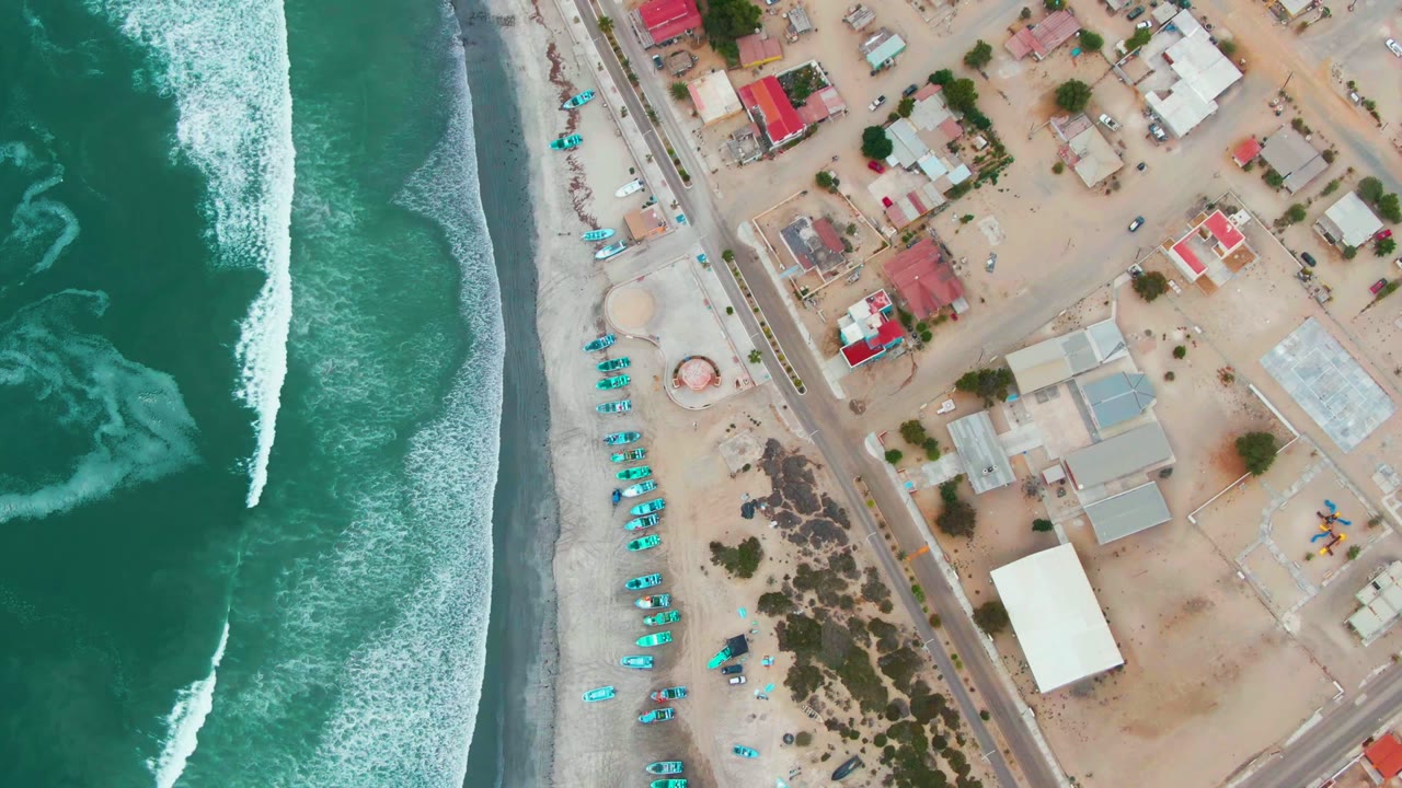 Top-down Punta Abreojos frame with white surf pushed closer to shore, a vertical line of blue umbrellas, a parallel road, scattered low buildings, and no visible people.