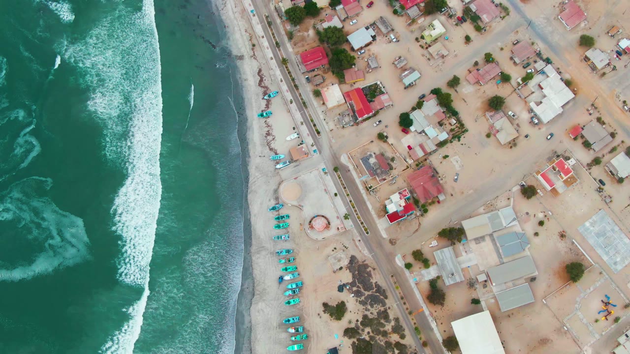 Overhead view in Punta Abreojos, Baja California Sur, with green-blue surf breaking along a sandy beach beside a coastal road, small boats, and homes, with no visible people.