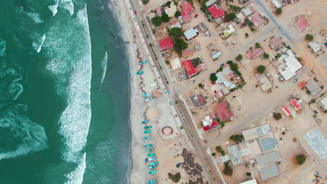 Drone-style overhead view in Punta Abreojos with textured green water, breaking white foam, a pale sandy beach, a parallel road, and a cluster of red-roofed and light-roofed homes, with no visible people.