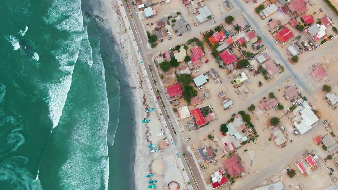 Overhead view in Punta Abreojos, Baja California Sur, with green ocean surf on the left, a sandy beach and road near center, residential homes on the right, and no visible people.
