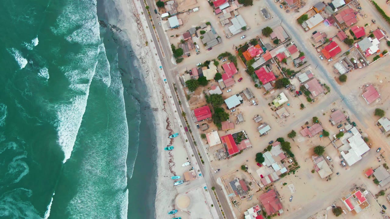 Drone view of white surf bands beside a beach road and residential blocks.