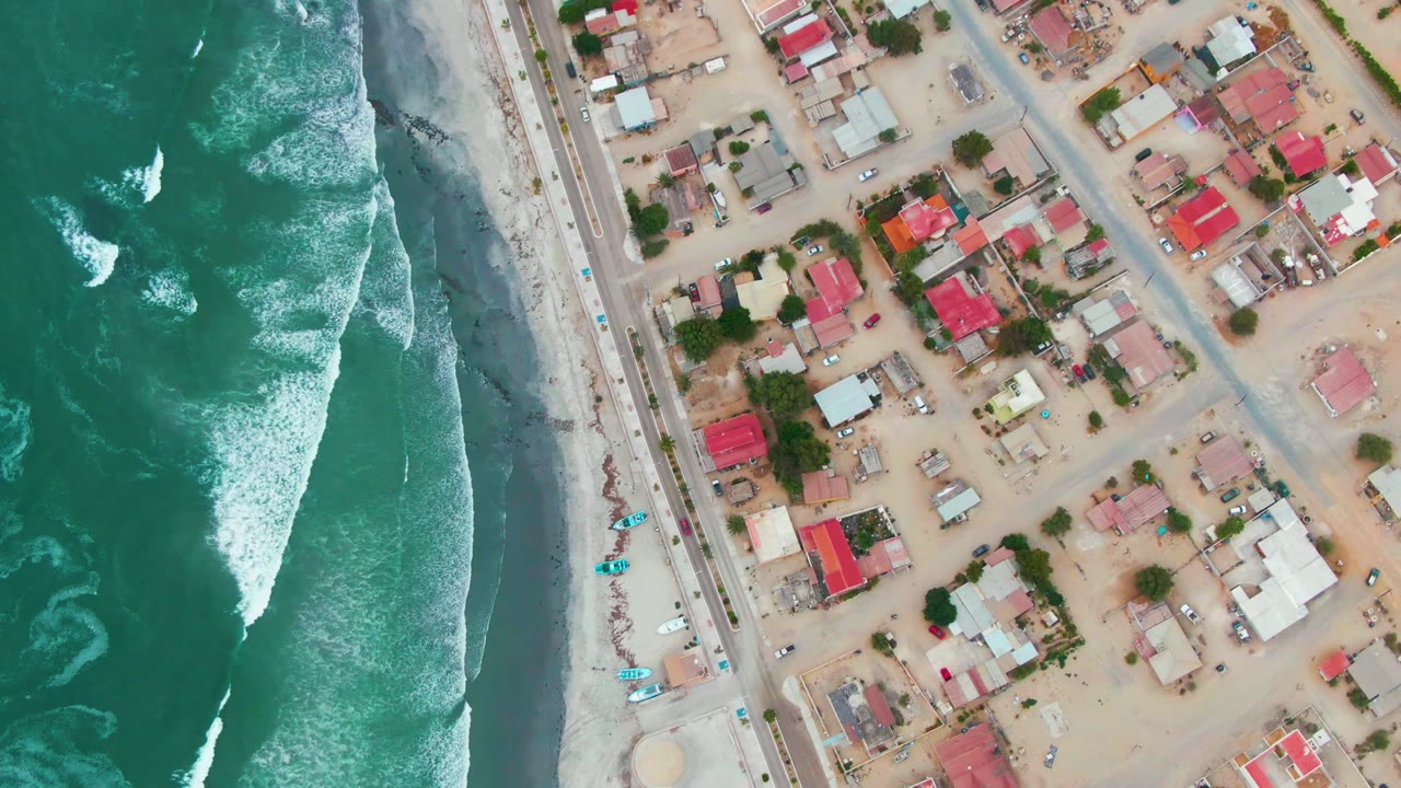 Overhead frame in Punta Abreojos with turquoise-green water and whitecaps at left, beach and road through the middle, and low residential buildings with red and light roofs at right, with no visible people.