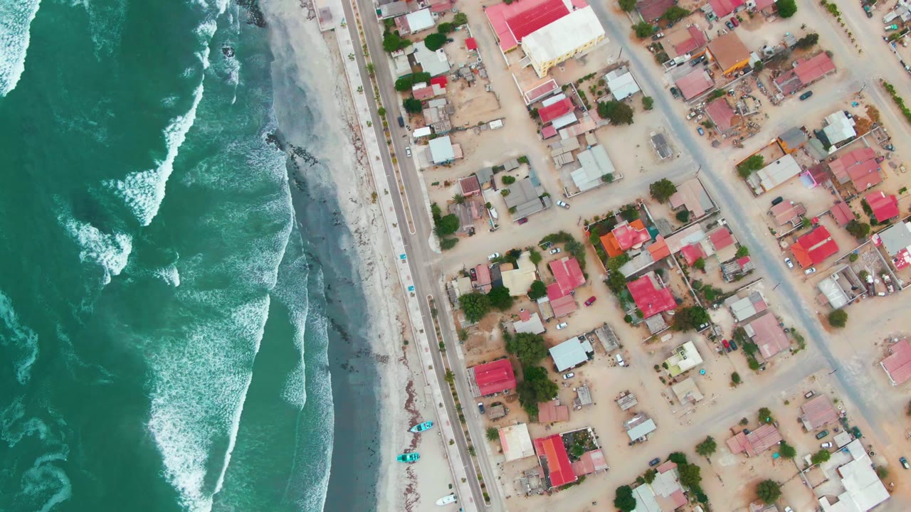 Aerial view of Punta Abreojos with green ocean surf on the left, a sandy beach and road in the center, and low residential buildings on the right, with no visible people.