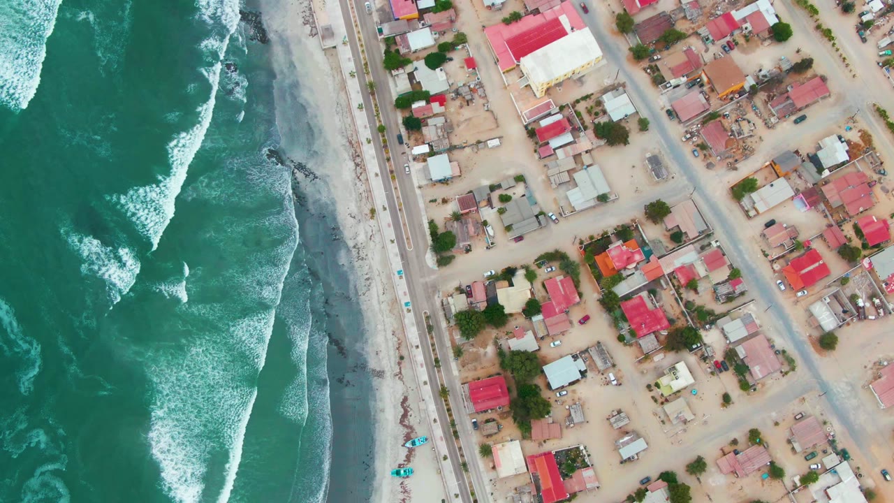 Aerial shoreline with breaking waves beside a road and town blocks.