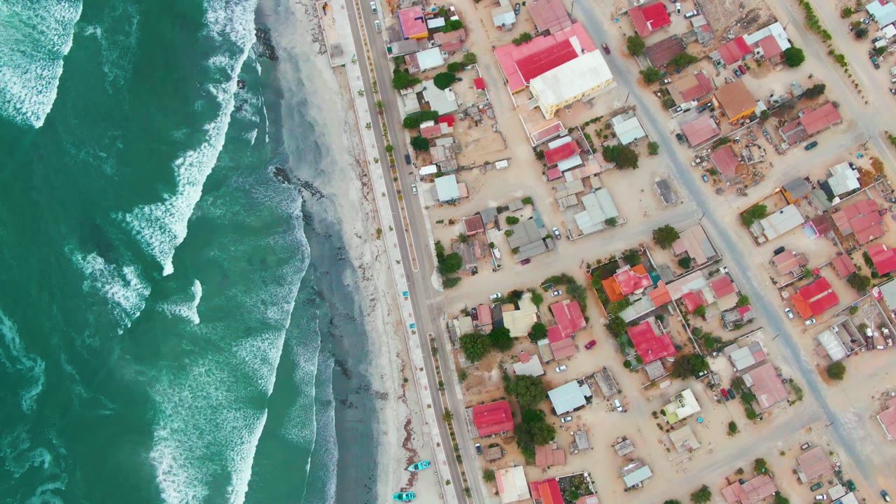 Overhead Punta Abreojos view with more white water visible offshore, a narrow beach strip, the coastal road, and dense low buildings inland, with no visible people.