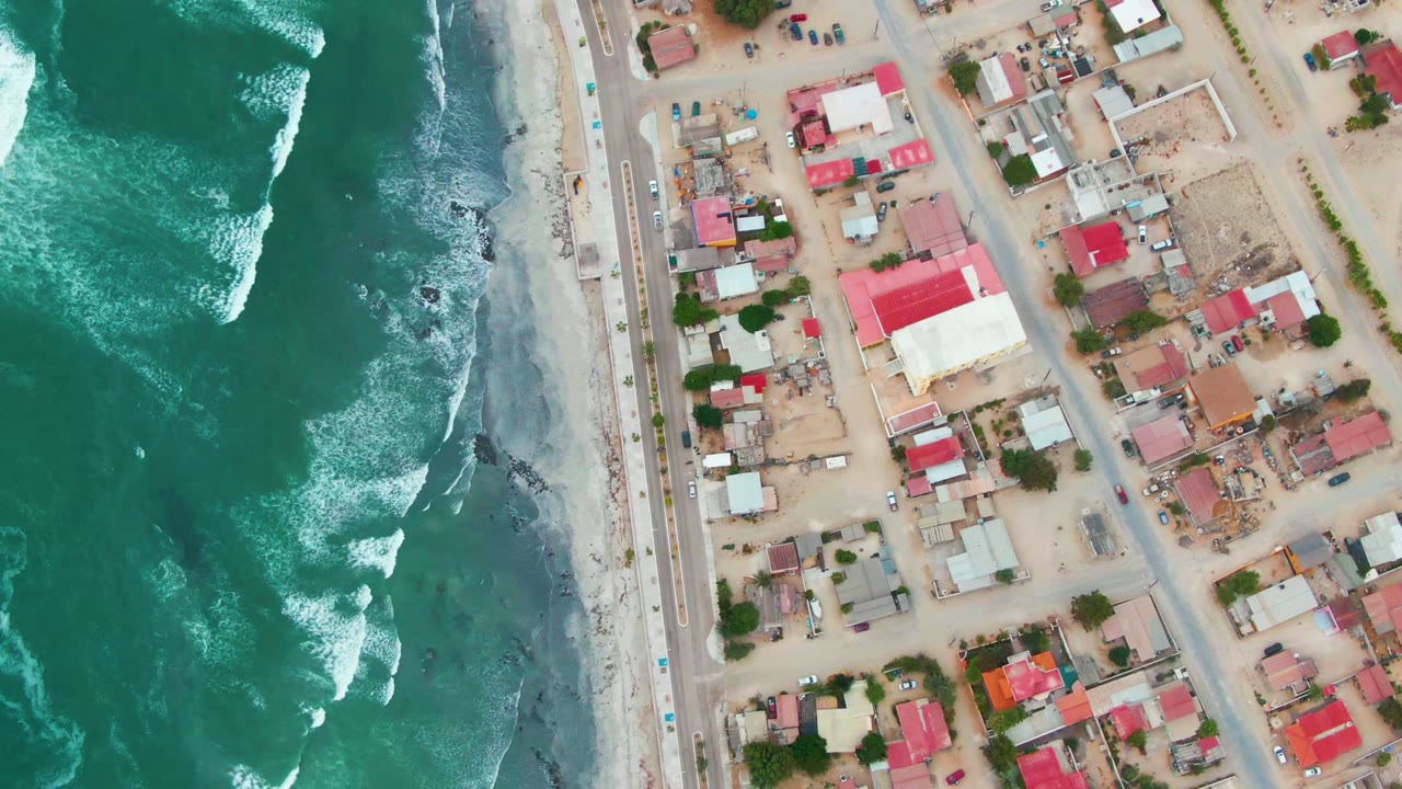 Opening frame over Punta Abreojos shows white surf breaking beside a narrow sandy beach, a straight coastal road, red-roofed buildings, sandy streets, and no visible people.