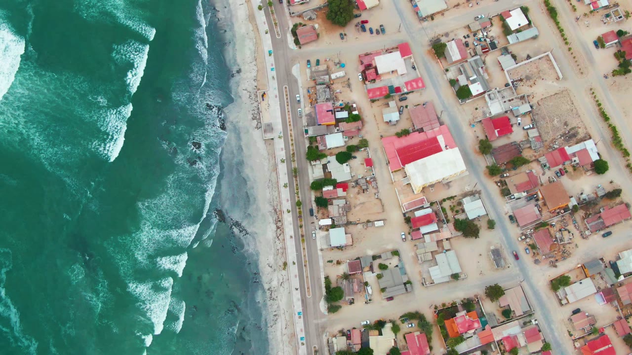 Aerial shoreline view with foamy waves, beach, road, and town blocks with red roofs.