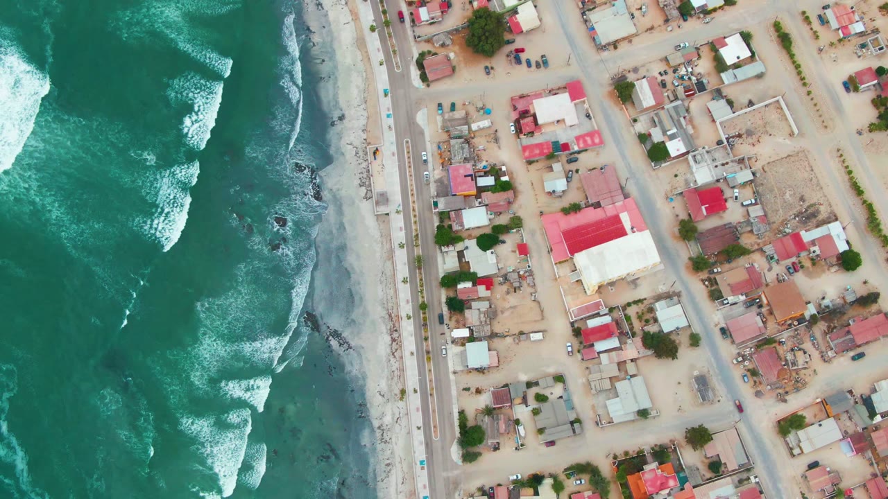 At two seconds, the overhead Punta Abreojos view shows a darker surf band, white breakers near shore, the road running parallel to the beach, red-roofed buildings, and no visible people.