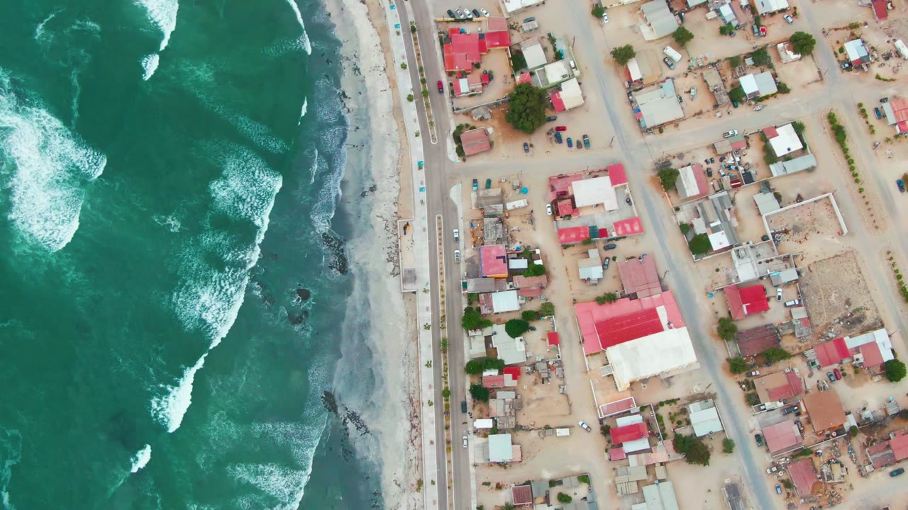 Overhead drone frame of Punta Abreojos, Baja California Sur, showing green ocean surf on the left, a sandy beach road, and low houses on the right with no visible people.