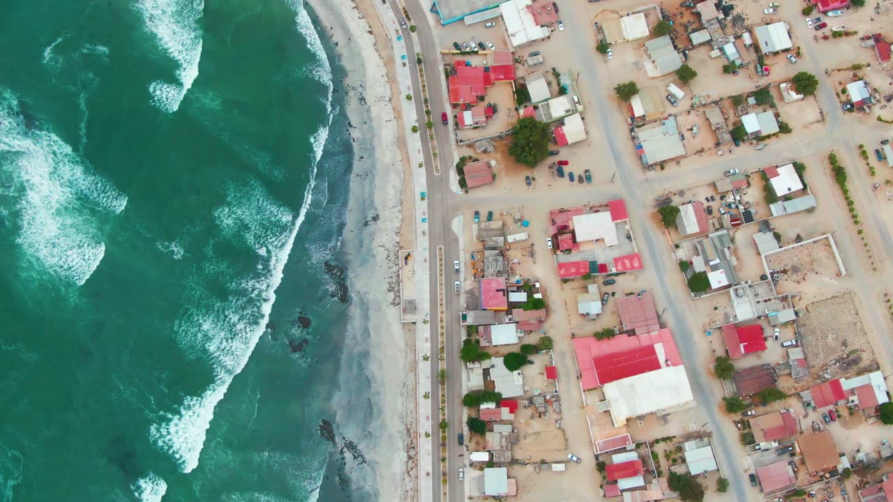 Top-down frame of Punta Abreojos coastline where darker green water and white foam meet the beach beside a road and residential lots, with no visible people.