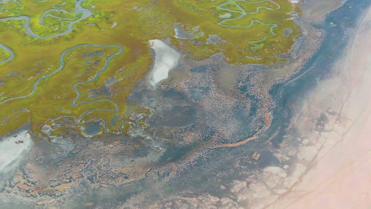 Aerial frame over Punta Abreojos wetland terrain with winding green marsh channels, dark tidal water, pale flats on the right, small white ice patches, and no visible people.