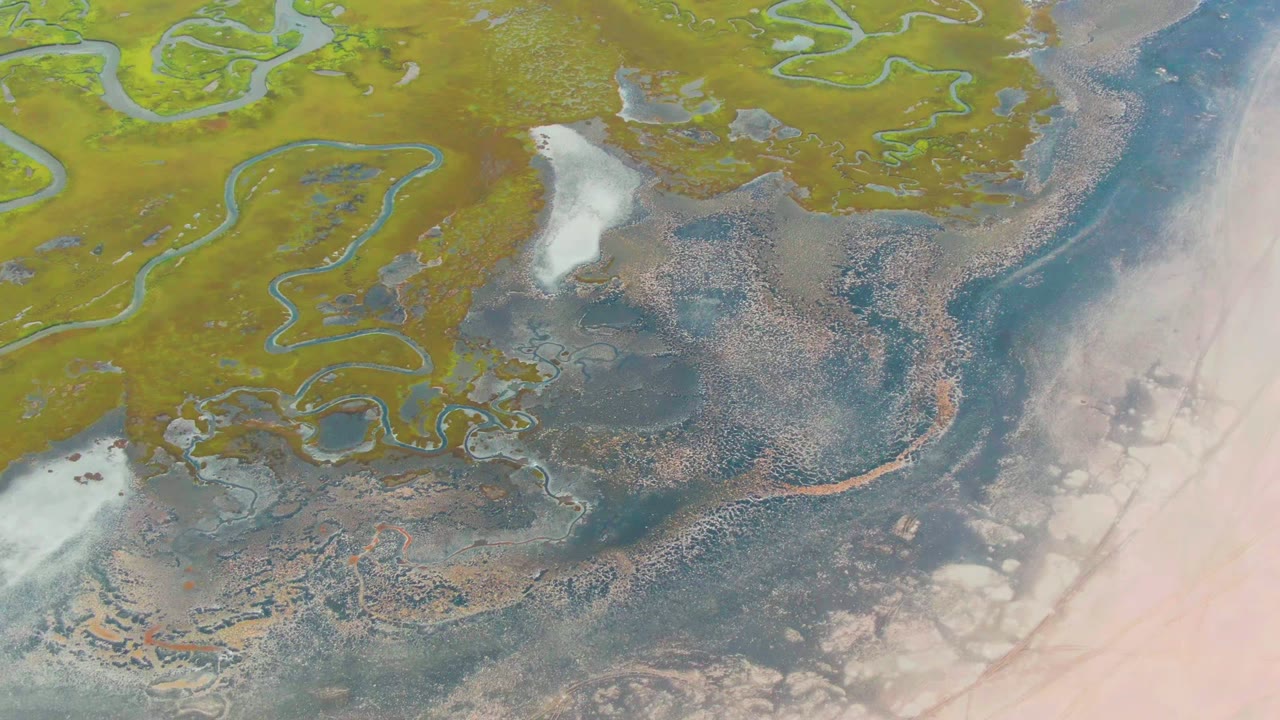 A high aerial frame over Punta Abreojos with green wetland channels at top left, brown thawed ground in the middle, dark blue water along the right, pale flats at the edge, and no visible people.