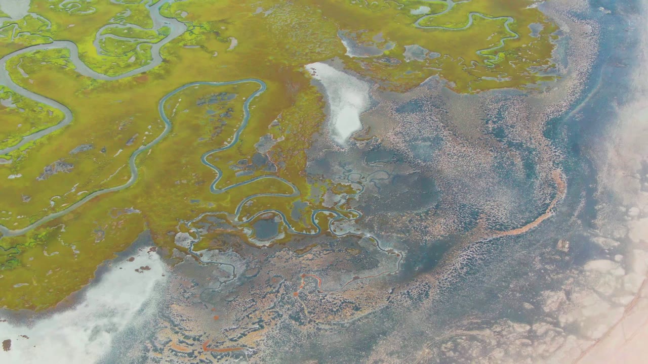 Aerial marsh channels with green land, wet flats, and small dark boats near water.