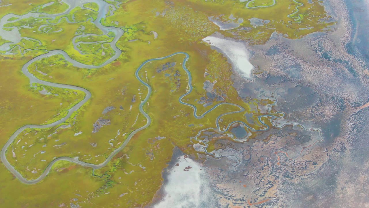 Overhead aerial frame from Punta Abreojos, Baja California Sur, showing winding pale blue water channels, green-brown terrain, white ice or snow patches, darker wet flats, and no visible people.
