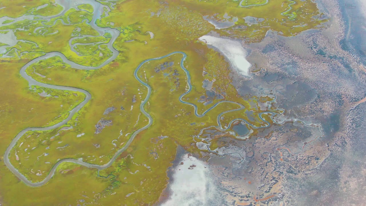 At two seconds, the overhead Punta Abreojos frame shows looping blue channels across green-yellow ground, mottled brown-gray wet areas to the right, a white ice or snow patch below center, and no visible people.
