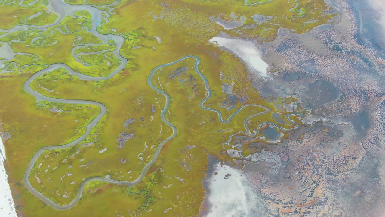 Aerial frame over Punta Abreojos showing winding streams through green and ocher wetland ground, pale flats near the shoreline, rippled blue-gray water, and no visible people.
