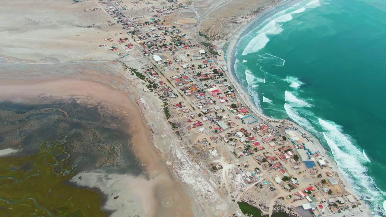 High aerial frame over Punta Abreojos, Baja California Sur, with turquoise ocean at right, white surf, a narrow beach road, compact buildings, pale inland sand, and no visible people.