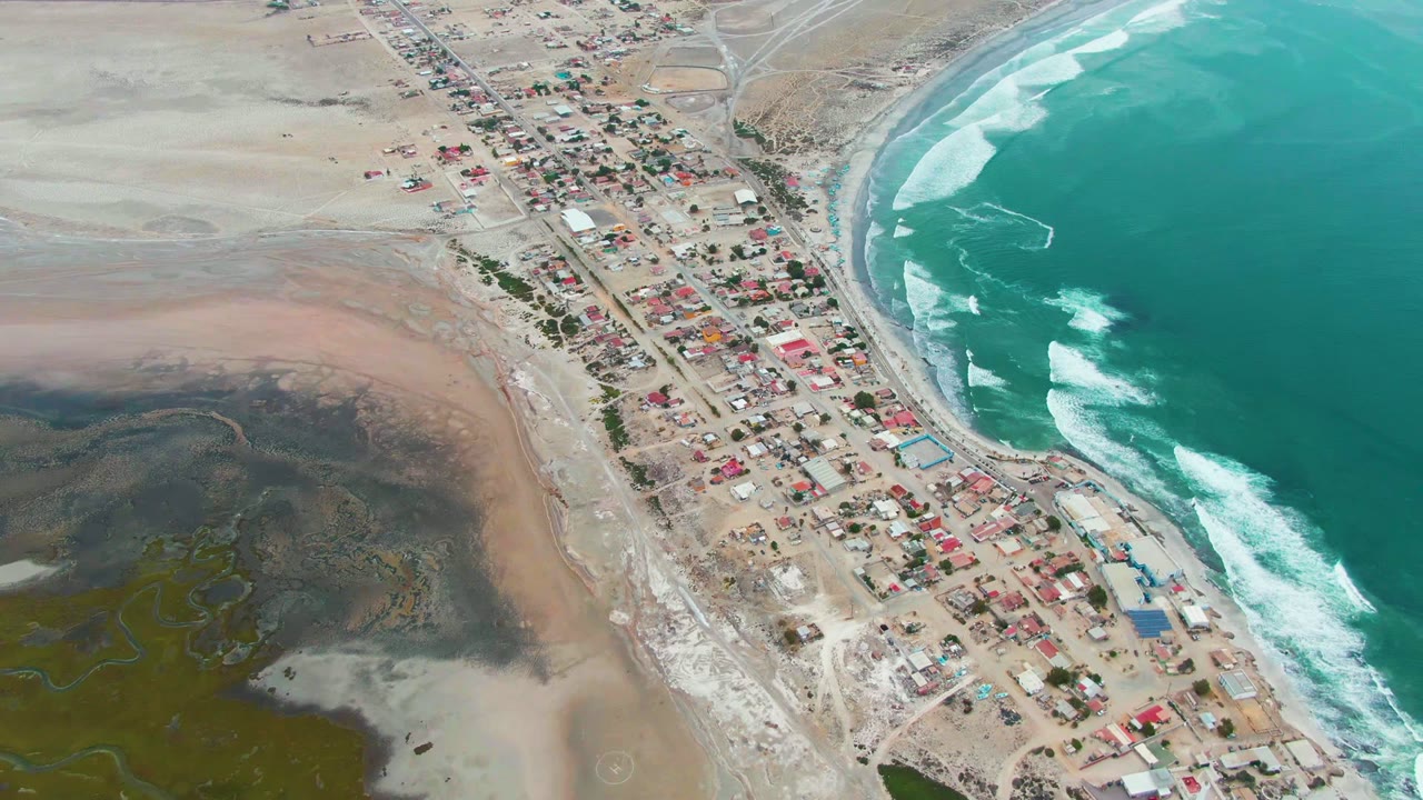 Coastal settlement running along a sandy road with ocean waves on the right.