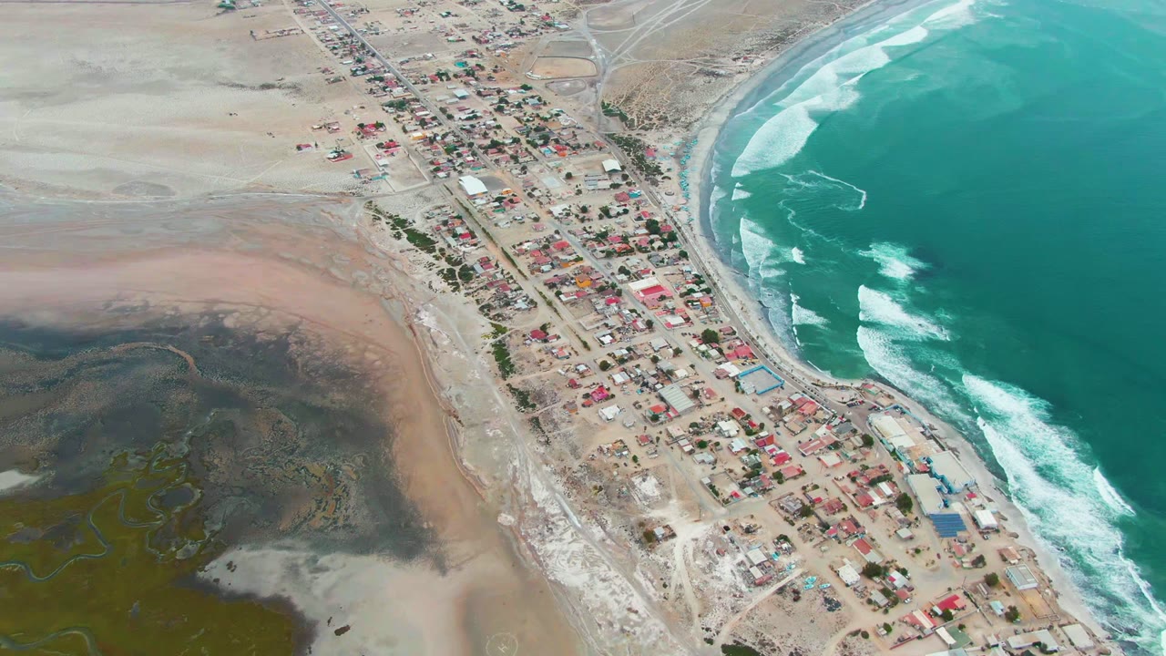 Punta Abreojos aerial frame with compact homes and structures near the beach, red roof accents, curling white surf on turquoise water, inland sand flats, and no visible people.