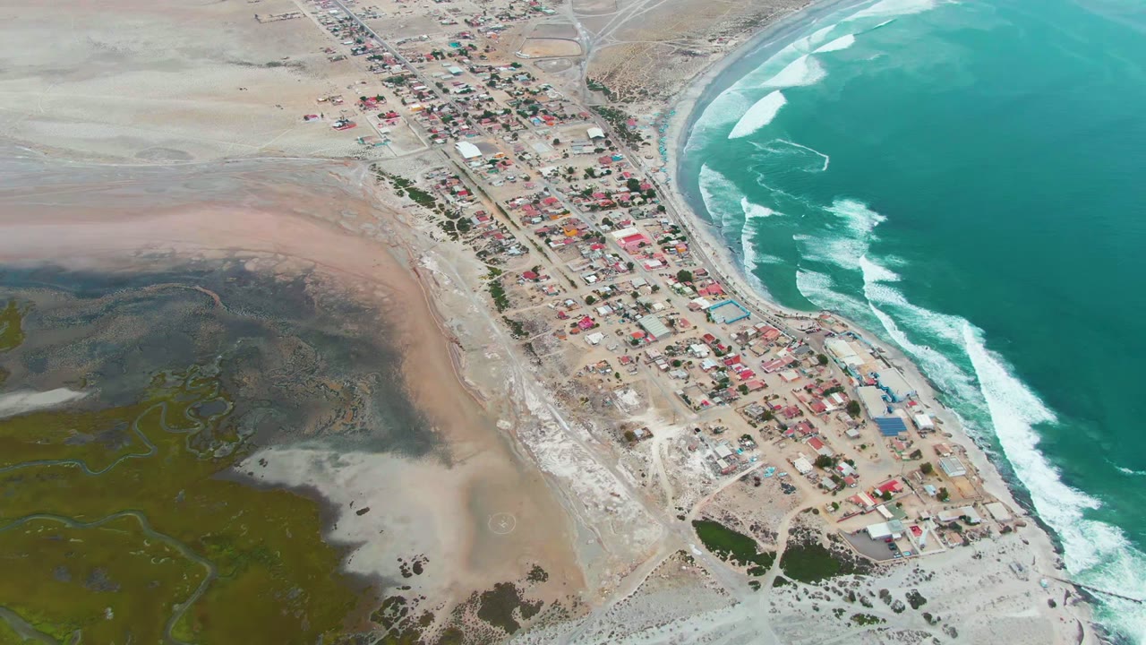 High aerial view over Punta Abreojos, Baja California Sur, showing a narrow coastal village beside turquoise surf and a green-brown estuary, with no visible people.